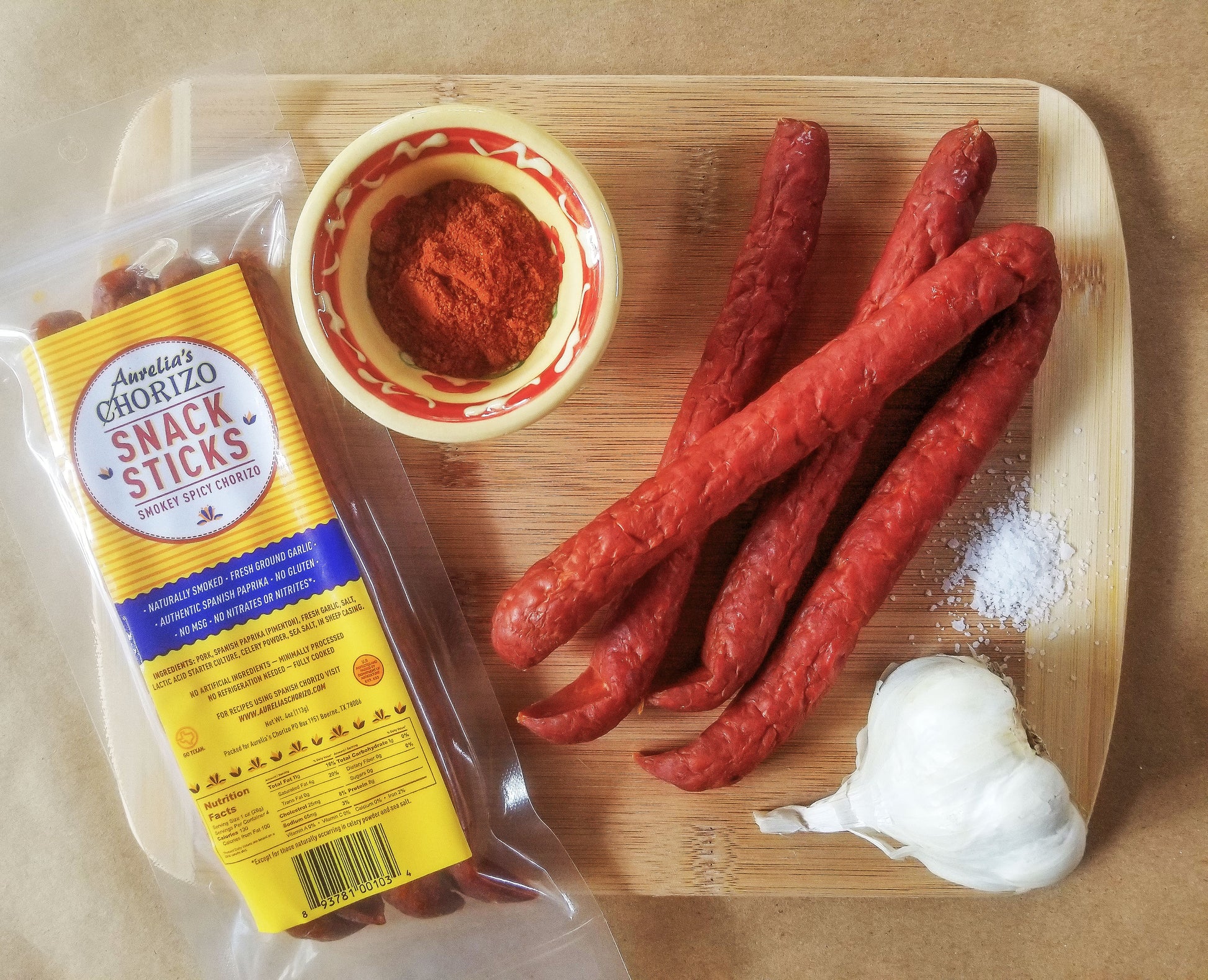 Cutting board with a package of Aurelia's Chorizo Snack Sticks along with a pinch of salt, a garlic clove, small dish of authentic Spanish Paprika and several Snack Sticks.